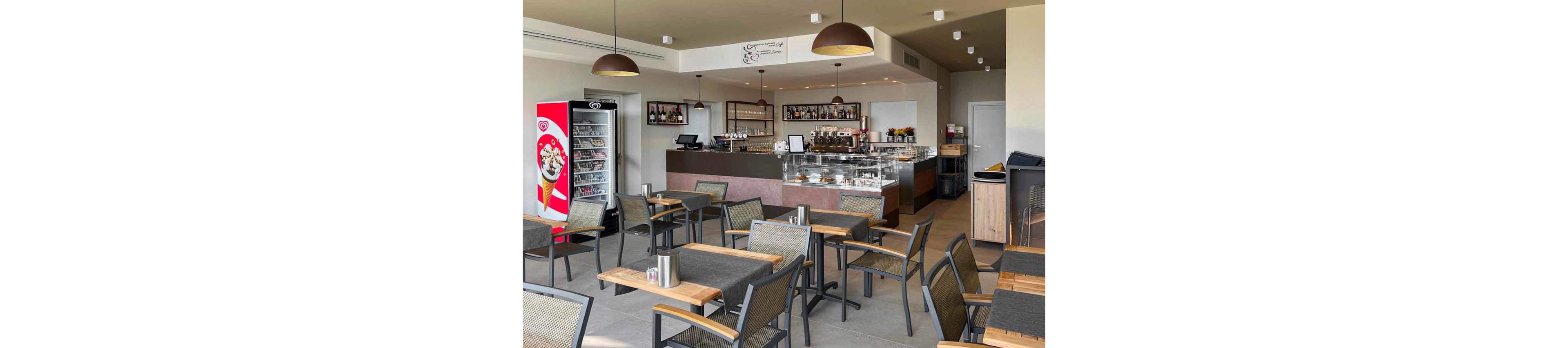 A contemporary café interior features neatly arranged tables and chairs facing a service counter. The counter area includes a glass display filled with desserts and a back bar with bottles and coffee equipment. On the left side, a tall refrigerated ice cream cabinet stands beside a snack fridge. Warm pendant lights and neutral colors create a welcoming atmosphere in the otherwise empty space.