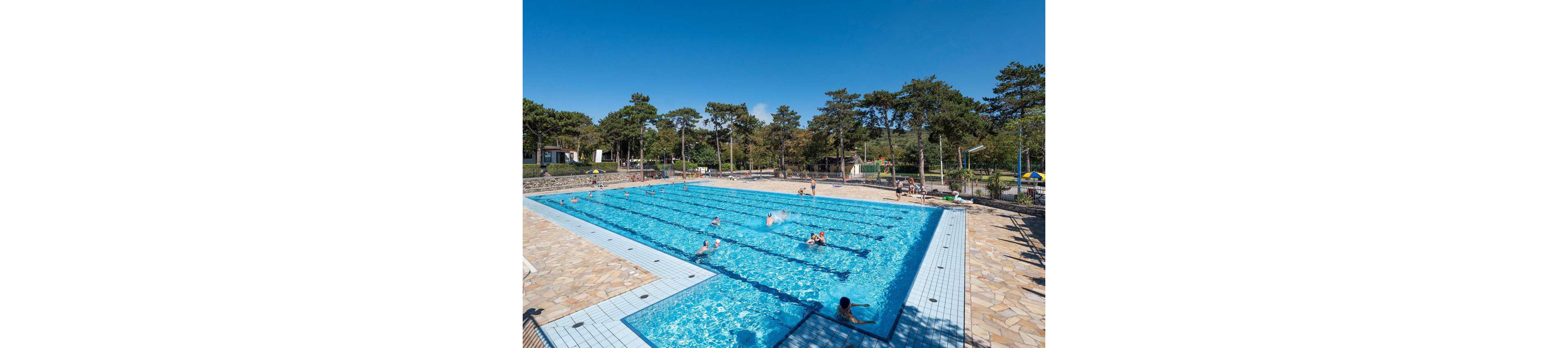 A large outdoor swimming pool is filled with people enjoying the water under a clear blue sky. The pool is divided into lanes and surrounded by a wide stone-tiled deck. Swimmers of different ages relax, play, and exercise in the bright sunlight. Tall trees and park buildings frame the scene, giving the area a recreational, family-friendly atmosphere.