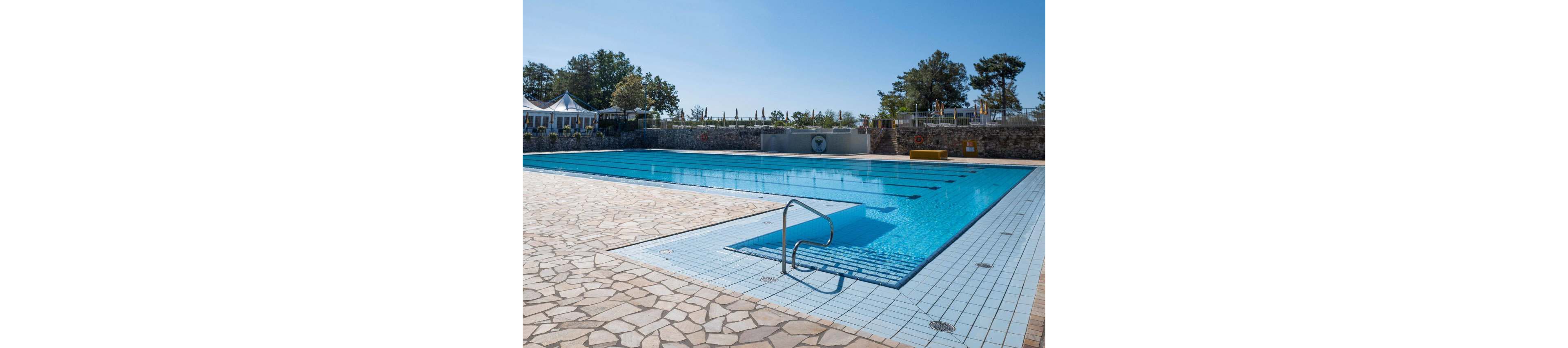 A large rectangular outdoor swimming pool sits empty of swimmers under a clear blue sky. The pool is surrounded by a wide stone-tiled deck with metal handrails leading into the water. In the background, a stone wall, trees, and a row of umbrellas and structures suggest a resort or recreational facility. Bright sunlight enhances the blue tones of the water and the relaxed, vacation-like atmosphere.