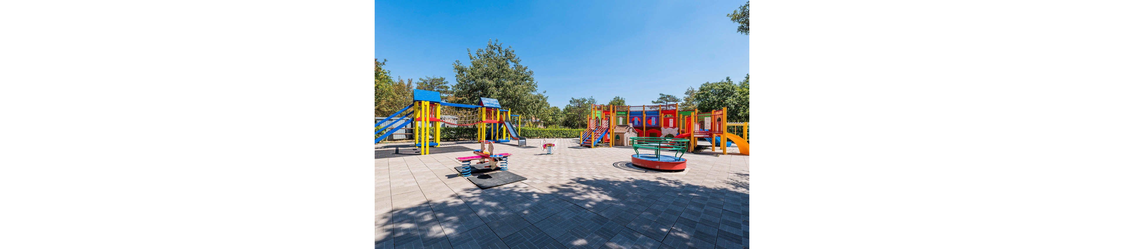 A bright, colorful playground is set in an open outdoor area on a sunny day. Multiple play structures with slides, climbing frames, and a merry-go-round are arranged on a tiled surface. The equipment is painted in vivid red, yellow, blue, and green tones, contrasting with the gray ground tiles. Mature trees and green hedges surround the playground, creating a safe and inviting recreational space for children.