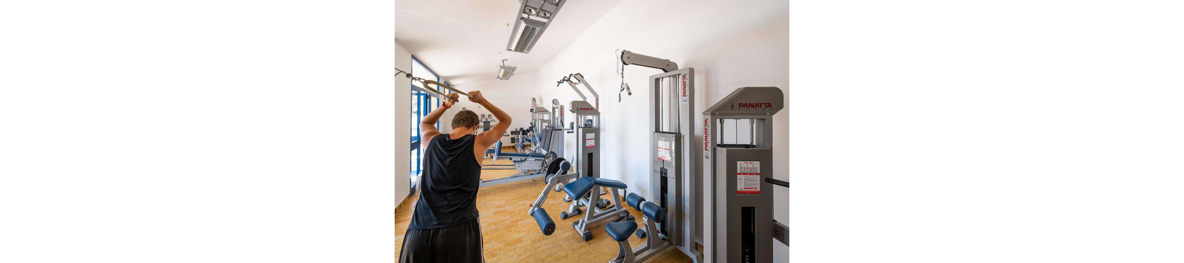 A young man exercises on a cable machine in a bright indoor gym. He is seen from behind wearing a sleeveless top while pulling the overhead bar. The room is filled with various strength-training machines, including seated and standing equipment. Large windows on the side wall bring in natural light, highlighting the clean, modern fitness space.