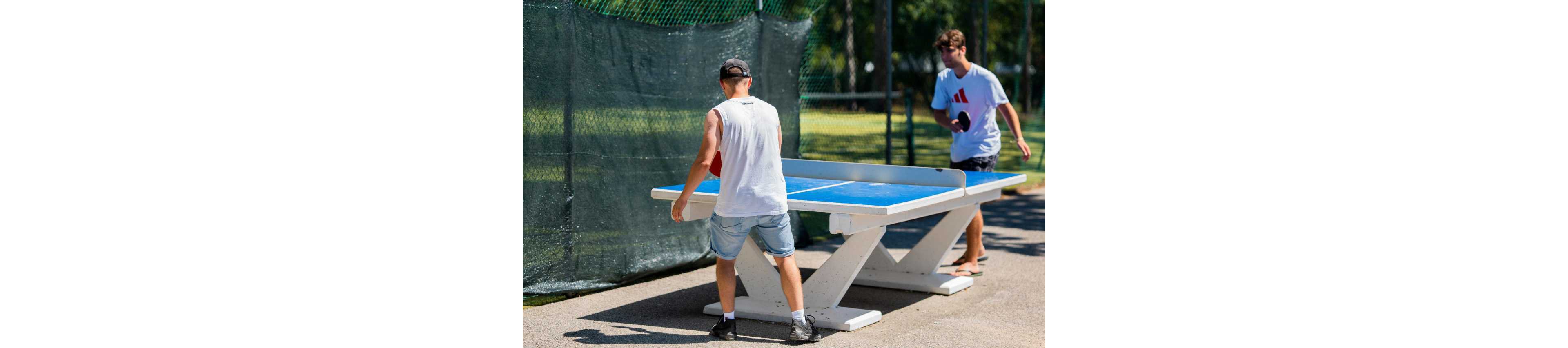 Two young men play table tennis on a blue outdoor table in a sunny park. One player in a white tank top and cap prepares to return the ball, while the other in a white T-shirt holds a paddle on the opposite side. A green mesh fence and trees create a casual recreational backdrop. The bright daylight and vivid colors emphasize an active, relaxed summer atmosphere.