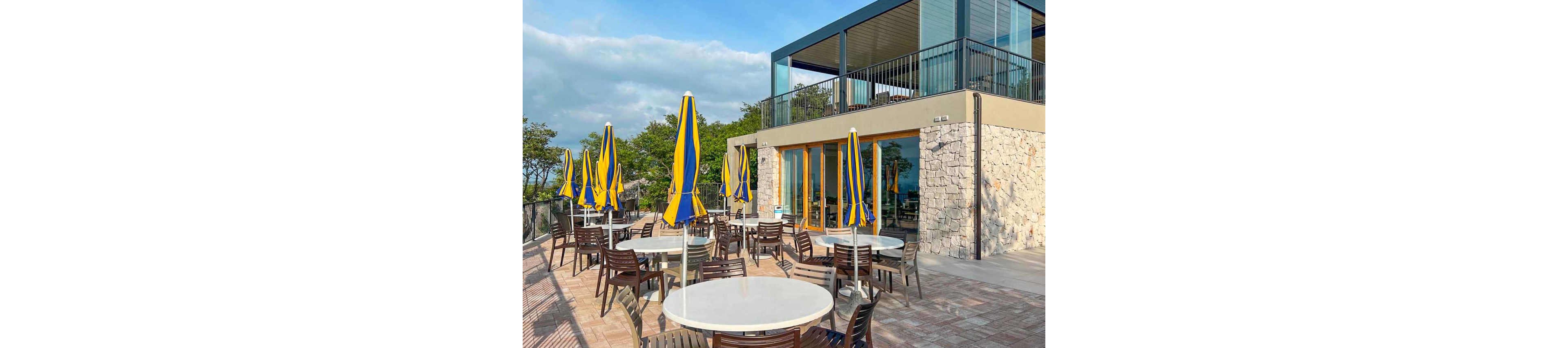 An outdoor café terrace features round white tables and numerous closed yellow and blue umbrellas. Brown chairs are neatly arranged around the tables on a paved patio. A modern two-story building with stone and glass walls borders the seating area, with trees and greenery in the background. The scene is brightly lit, suggesting a pleasant, sunny day with partly cloudy skies.