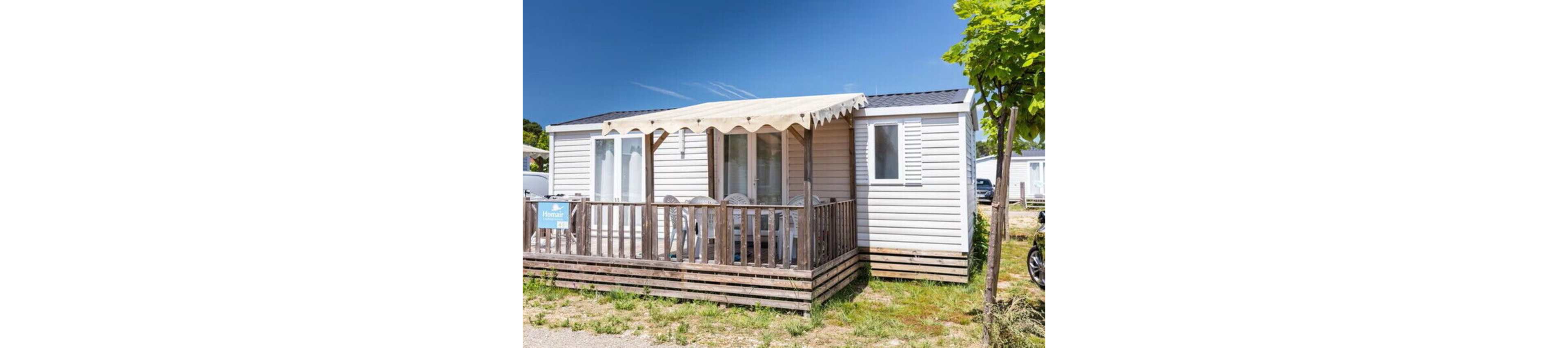 A compact mobile home with white siding stands under a clear blue sky. The front features a wooden terrace with railing and a light-colored awning providing shade. Several white plastic chairs and a table are arranged on the deck, suggesting an outdoor relaxation area. The setting appears to be a holiday park or campground with other similar units visible in the background and a small tree in the foreground.
