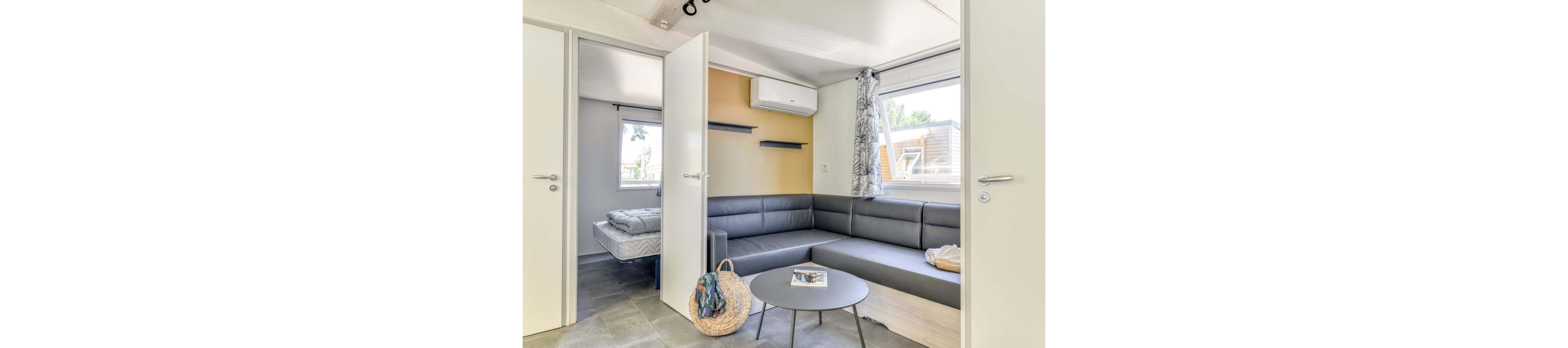 Bright living area inside a modern mobile home features a gray sectional sofa and round coffee table. Open doors reveal a bedroom with a neatly made bed, emphasizing the compact yet functional layout. Large windows with patterned curtains bring in natural light and views of neighboring cabins. A woven beach bag and magazine on the table suggest a relaxed vacation atmosphere.