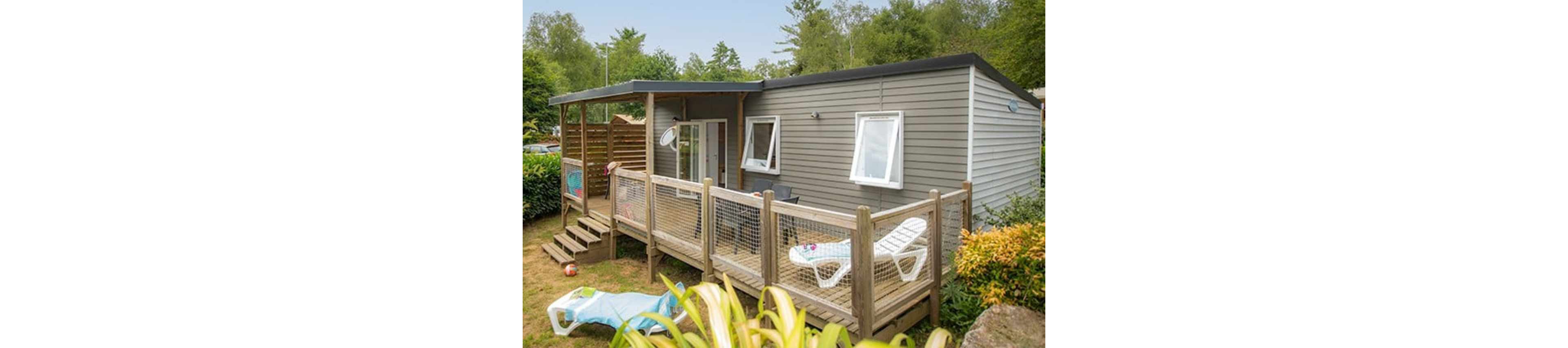 A modern mobile home with gray siding features a spacious wooden deck surrounded by a simple railing. White lounge chairs and a small table are arranged on the deck, suggesting a relaxing outdoor space. The home is set in a lush, green garden with trees and shrubs providing a natural backdrop. A child's inflatable pool toy and scattered outdoor items add a casual, family-friendly atmosphere.