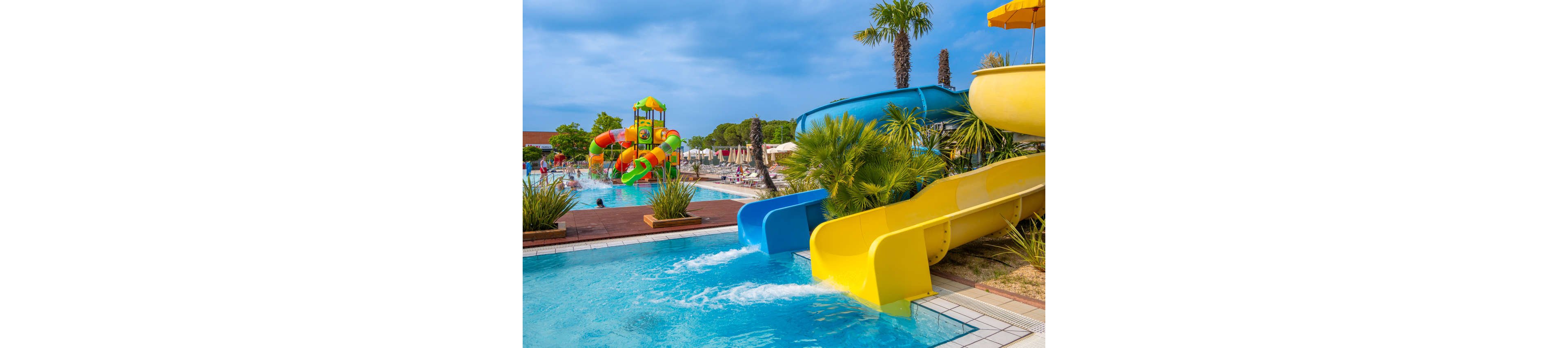 A vibrant outdoor water park features bright yellow and blue slides emptying into a clear swimming pool. In the background, families enjoy additional colorful water play structures and shallow pools. Palm trees and lush greenery surround the pool area, creating a tropical resort atmosphere. The sky is partly cloudy, suggesting a warm and pleasant day for leisure and fun.