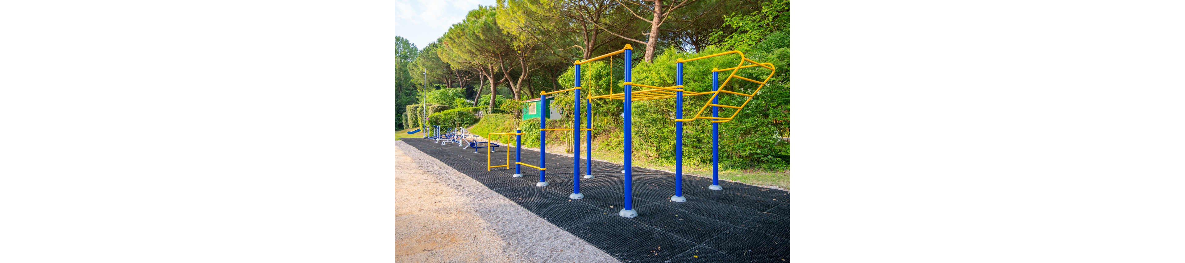 An outdoor fitness area features bright yellow and blue calisthenics bars on a black rubber surface. The equipment is set along a path surrounded by lush green trees and bushes. Additional exercise machines are visible further down the track, creating a full workout circuit. The scene suggests a public park designed for bodyweight training and recreational exercise.