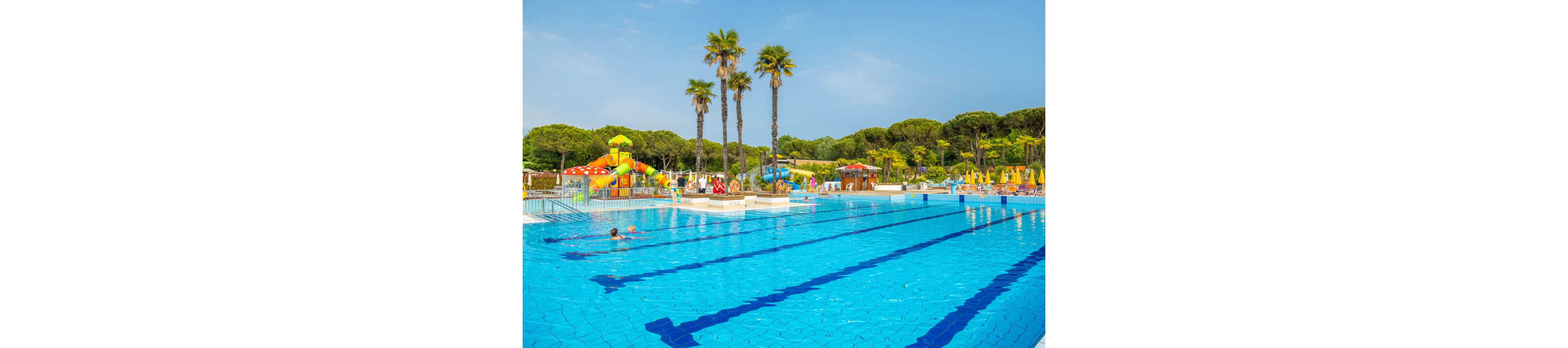 Large blue outdoor swimming pools stretch across the foreground under a clear sunny sky. A few people relax and swim in the water while others enjoy the surrounding deck. In the background, colorful water slides and play structures rise near rows of yellow sun loungers and umbrellas. Tall palm trees and dense green forest create a lush holiday atmosphere.