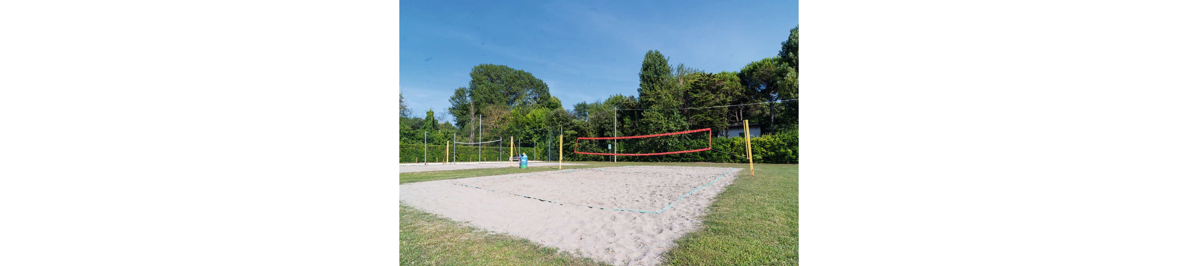 An empty sand volleyball court is set up in an outdoor park. The court is surrounded by green grass, trees, and shrubs under a clear blue sky. Yellow poles and a red net define the main court, with additional courts visible in the background. The scene suggests a quiet, sunny day suitable for recreational sports and leisure activities.