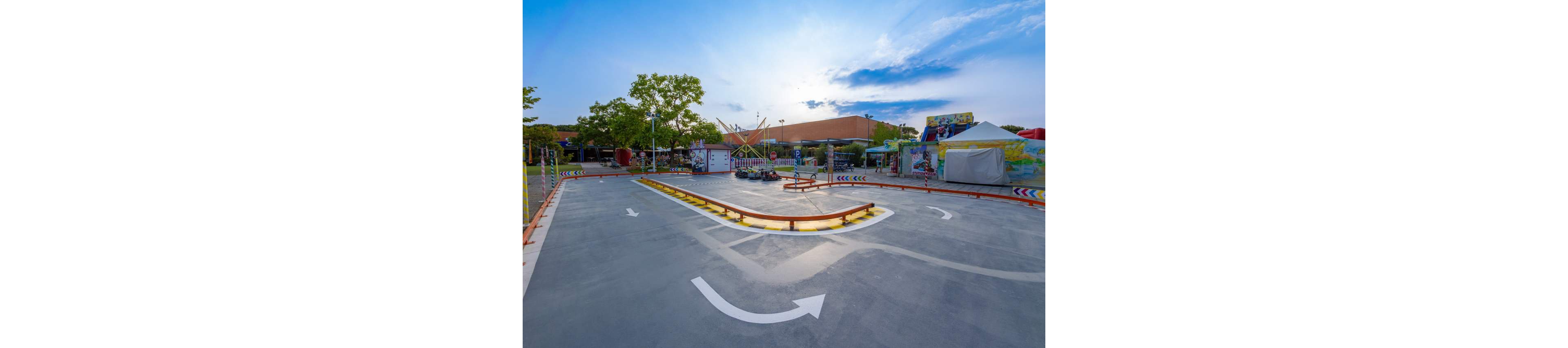 An empty go-kart track is shown in an outdoor amusement park under a bright blue sky. The track features painted white directional arrows and colorful striped barriers guiding the driving path. In the background are trees, tents, and various fairground structures, suggesting a family entertainment area. The warm evening light and clear weather create a calm, inviting atmosphere.