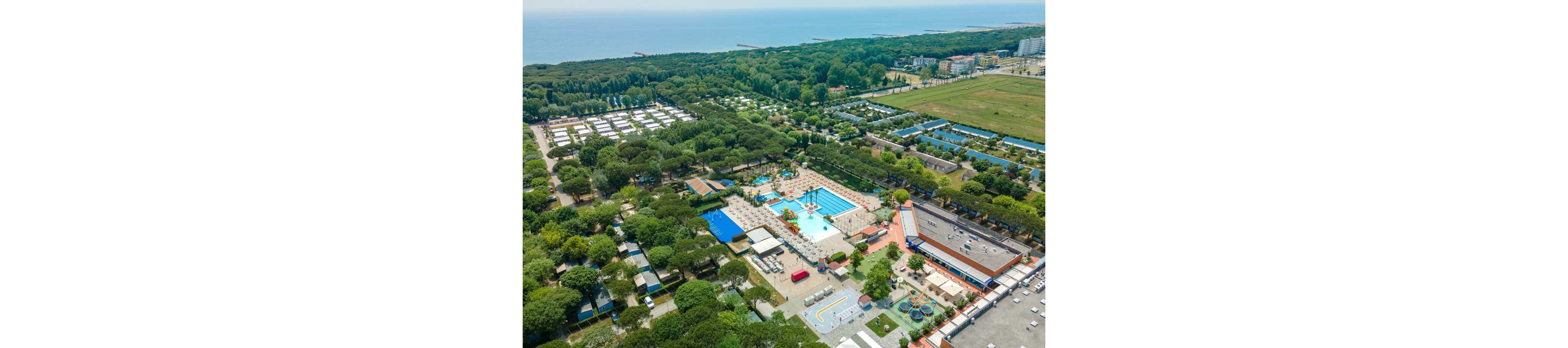 A large holiday resort complex is seen from above, surrounded by dense green trees near a coastline. Multiple outdoor swimming pools with slides and sun loungers form the central focus of the property. Nearby are camping or bungalow areas, sports courts, and children’s play zones. In the distance, the sea and sandy beach stretch along the horizon, emphasizing a relaxed vacation setting.