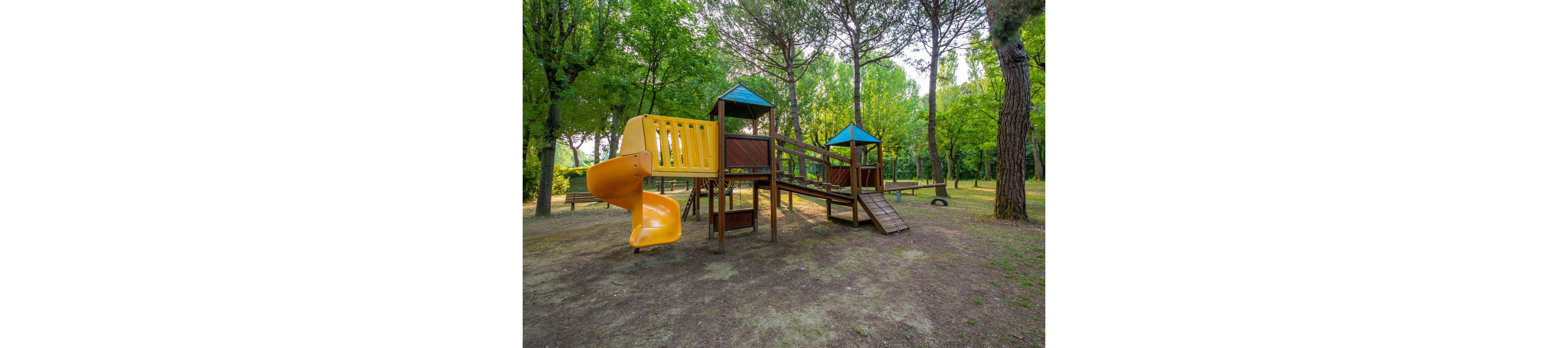 A wooden playground structure with a bright yellow slide stands in a quiet park. The play area is surrounded by tall green trees and dappled sunlight on the ground. Ramps, platforms, and a small bridge connect different parts of the structure. No children are present, emphasizing the peaceful, natural setting.