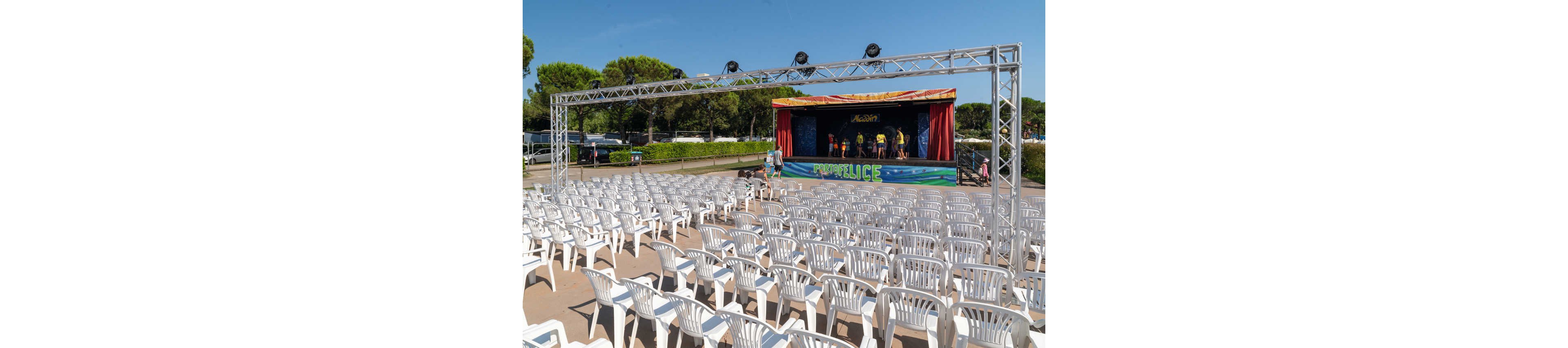 An outdoor performance stage is set up in front of rows of empty white plastic chairs. A few people stand near the brightly colored stage where performers are visible under a red and blue canopy. A large metal truss frame spans above the seating area, suggesting lighting or sound equipment support. The scene is surrounded by green trees and hedges under a clear blue sky, indicating a sunny day event setting.