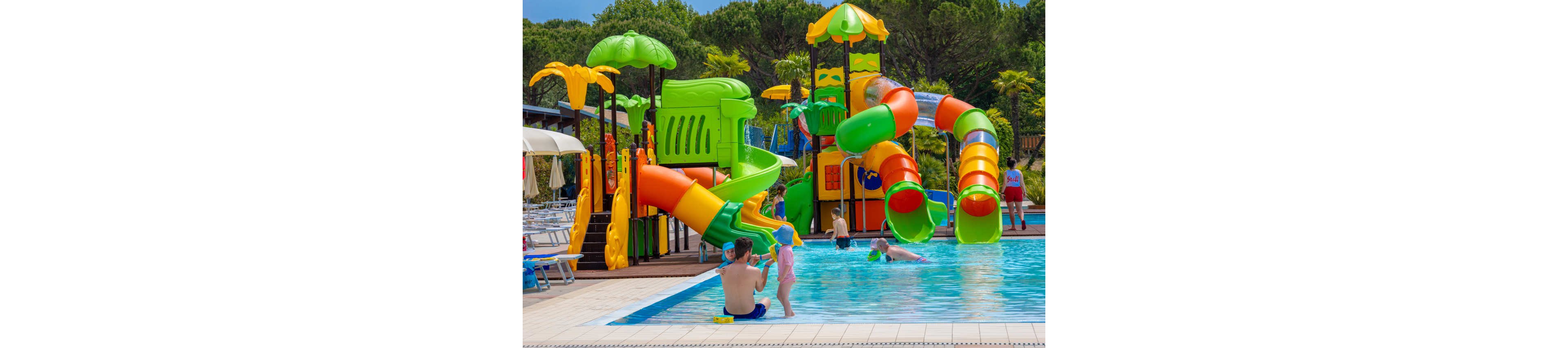 A vibrant outdoor water park features bright green, orange, and yellow slides and climbing structures beside a swimming pool. Several children and adults play and relax in the shallow water near the edge of the pool. The scene is set on a sunny day with lush green trees in the background and lounge chairs arranged along the deck. The overall atmosphere is fun, family-friendly, and summery.
