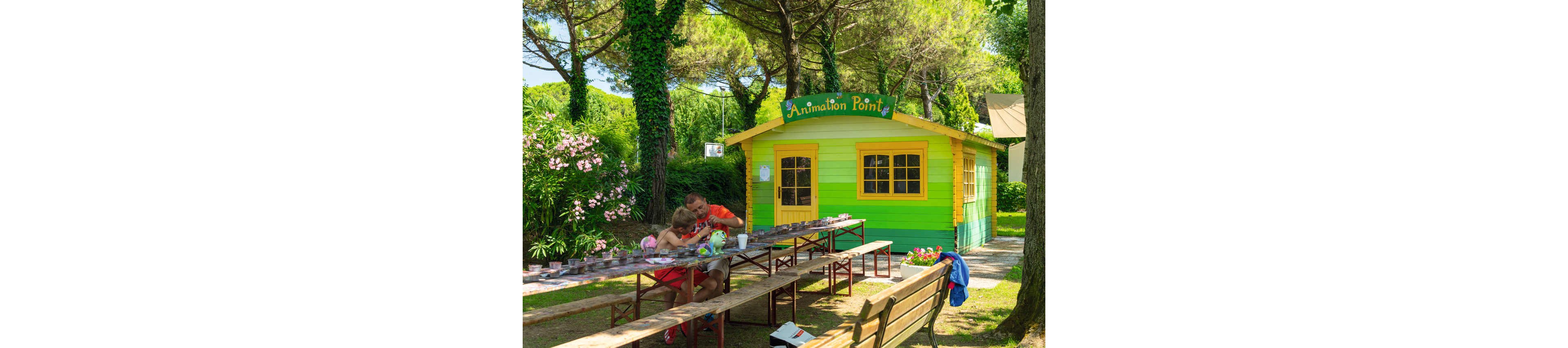 A father and child sit together at long outdoor tables working on crafts in front of a small colorful hut. The wooden building is painted in bright yellow and green with a sign reading "Animation Point" above the door. The scene is set in a lush garden with tall trees, green foliage, and blooming flowers surrounding the area. Sunlight and vibrant colors create a cheerful, relaxed holiday atmosphere.