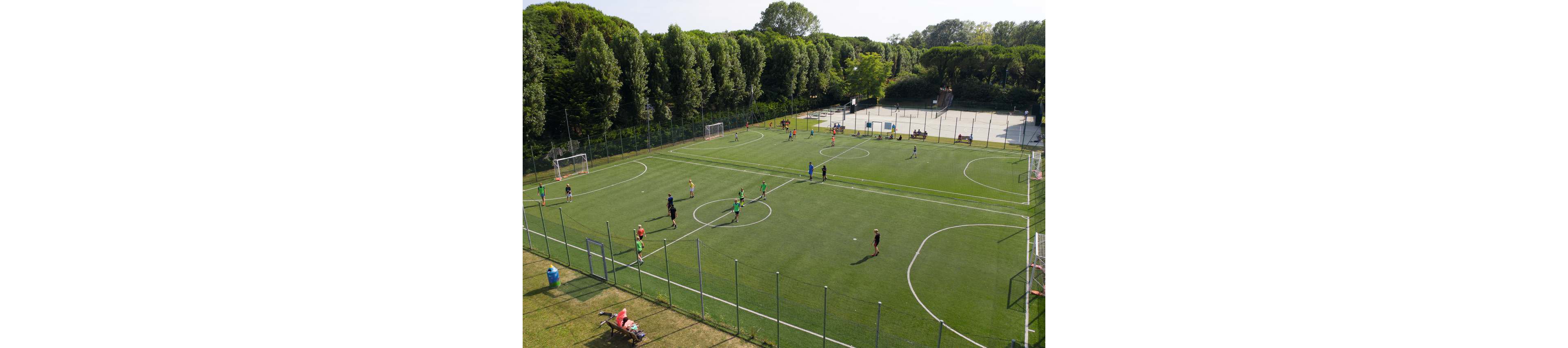 An amateur soccer game is being played on a large outdoor artificial turf field. Players in colorful jerseys are spread across the pitch, with a few people watching from the sidelines. The field is surrounded by tall green trees and enclosed by a wire fence. In the background, additional sports courts and open recreational areas are visible under clear daylight.
