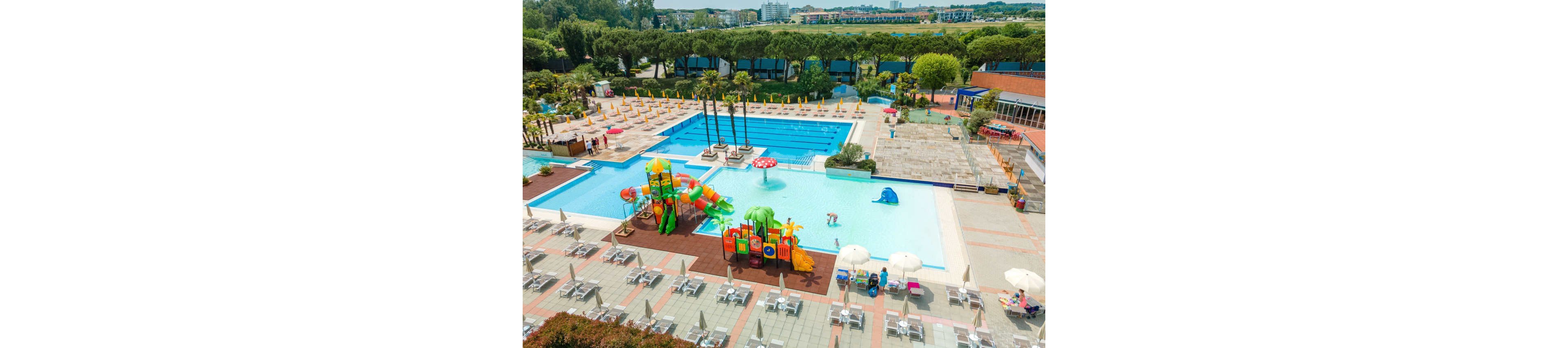 A large outdoor swimming pool complex features multiple pools and a colorful children’s water play structure. Sun loungers and umbrellas are neatly arranged around the pools, suggesting a resort or holiday setting. A few people relax and play in the shallow water near the slides and fountains. Tall trees and distant buildings frame the background, creating a sunny, inviting vacation atmosphere.