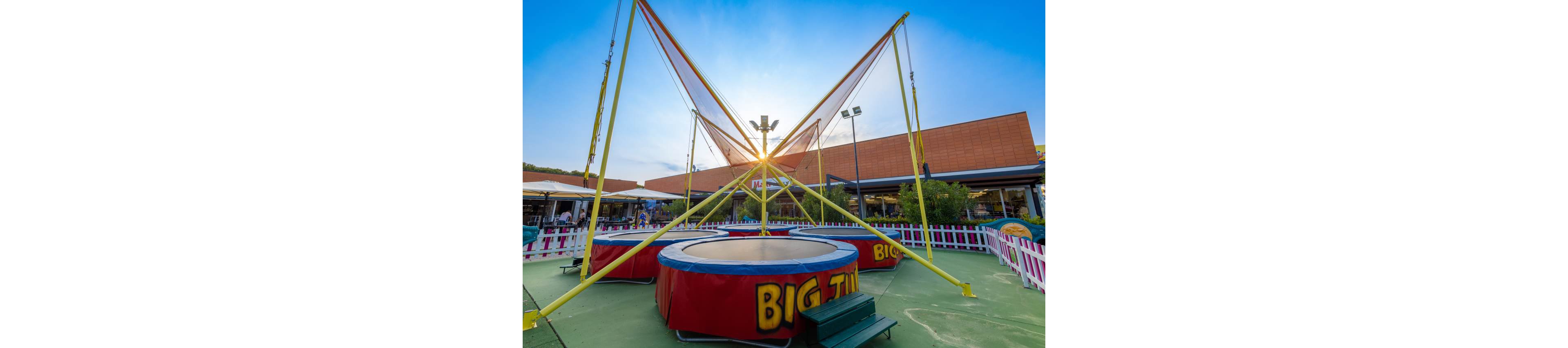 An outdoor bungee trampoline attraction stands in the center of a fenced play area. The structure features tall yellow poles and red padded bases with the words "BIG JUMP" clearly visible. A shopping or entertainment complex with brick facades and storefronts forms the background. The sun sets behind the structure, creating a bright sky and long shadows across the ground.