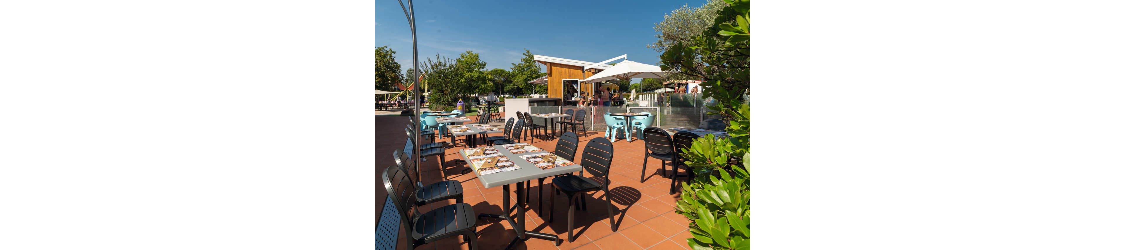 An outdoor café terrace features several empty tables and black chairs arranged on a terracotta-tiled floor. Menus are laid out on the tables, ready for guests. In the background, a small modern kiosk or bar with umbrellas provides shade for additional seating. Green shrubs and trees surround the area, creating a relaxed, summery atmosphere under a clear blue sky.