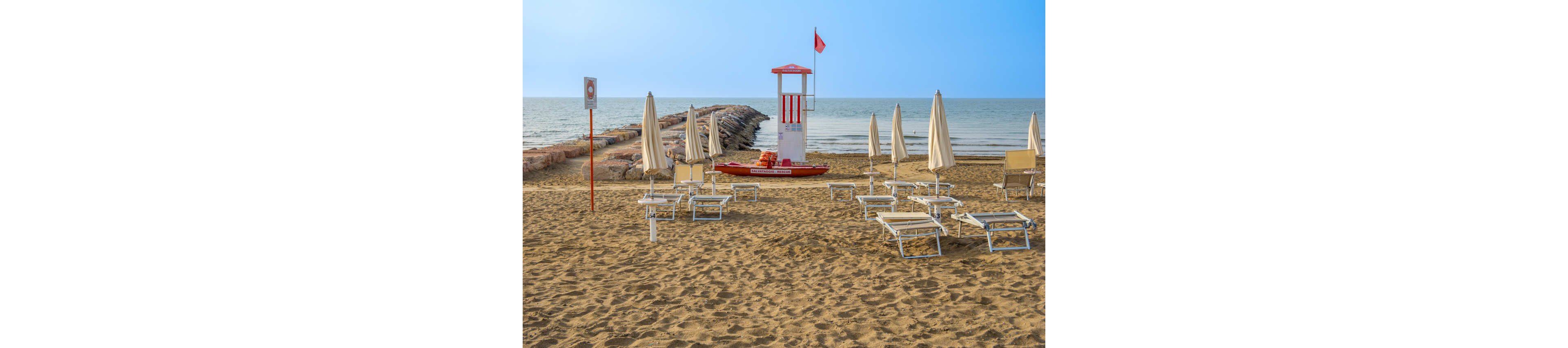 A quiet sandy beach is lined with empty lounge chairs and closed umbrellas facing the sea. In the center stands a white and red lifeguard tower with a small rescue boat at its base. A rocky breakwater extends into the calm water under a clear blue sky. The scene feels peaceful and off-season, with no people present on the beach.
