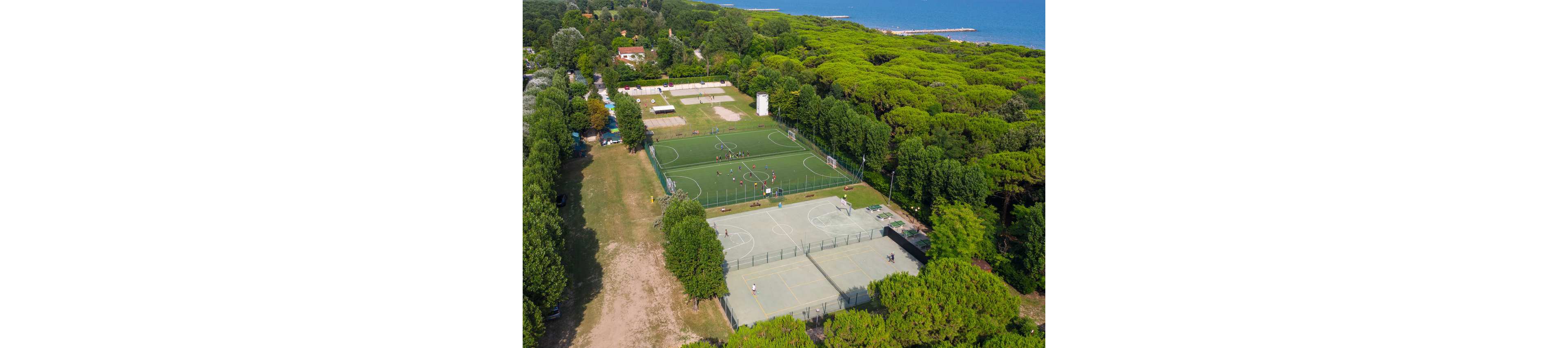A drone captures an aerial view of multiple outdoor sports courts surrounded by dense green forest near a coastline. In the center, people play on a green artificial turf soccer field enclosed by fencing. Adjacent light-colored courts appear suitable for tennis or other court sports, with clear boundary lines and nets. The scene is bright and sunny, with nearby buildings and pathways visible among the trees, creating a recreational park atmosphere.
