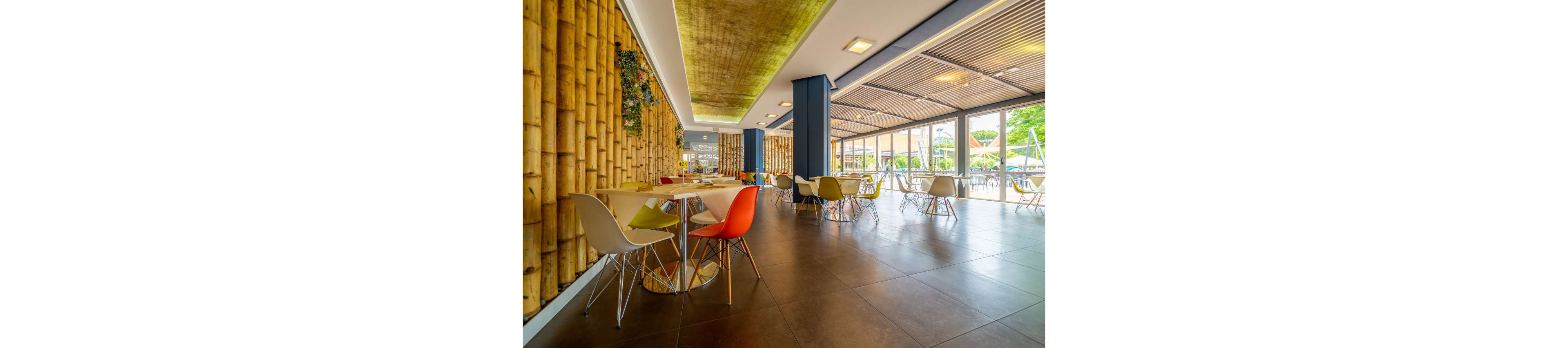 A bright, modern cafe interior features colorful chairs around light wooden tables. Large floor-to-ceiling windows open to an outdoor area with greenery and playground equipment. One wall is decorated with vertical bamboo, adding a natural accent to the contemporary design. Warm lighting and spacious seating create an inviting, relaxed atmosphere.