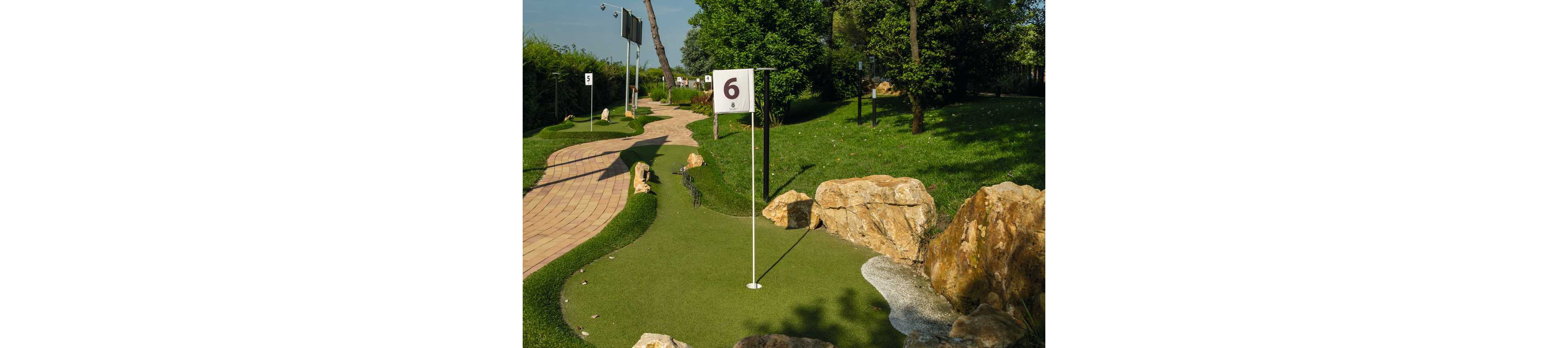 An outdoor mini golf course features a green with a flag marked with the number 6. The putting lane winds between large rocks and landscaped grass under bright sunlight. A brick pathway runs alongside the course, leading past additional numbered holes in the background. Trees and shrubs surround the area, creating a relaxed recreational setting.