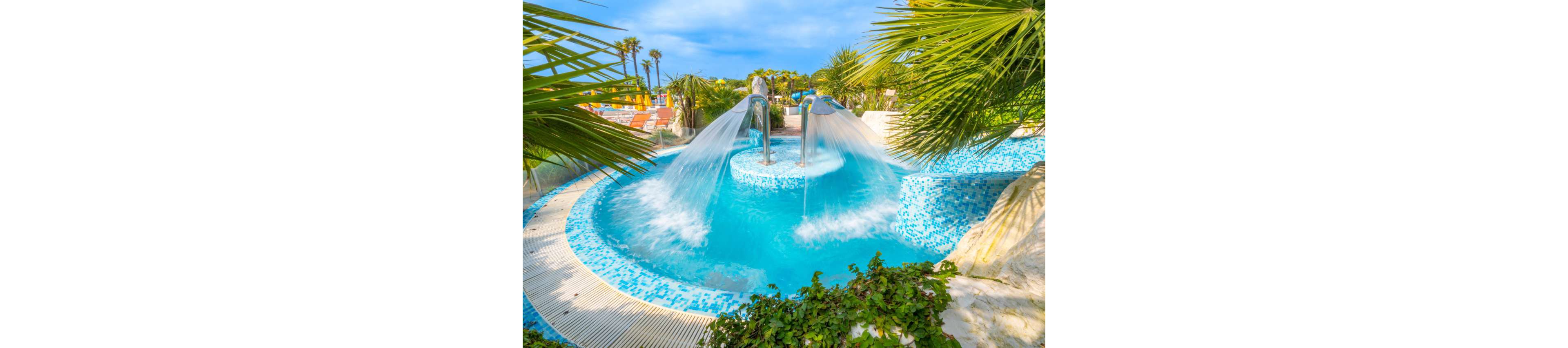 An outdoor resort-style pool features strong cascading water streams flowing from metal spouts into a bright blue basin. The pool is tiled with a mosaic of light and dark blue tiles, creating a vibrant aquatic look. Palm trees and sun loungers are visible in the background, suggesting a warm vacation setting. Lush green plants frame the foreground, enhancing the tropical and relaxing atmosphere.