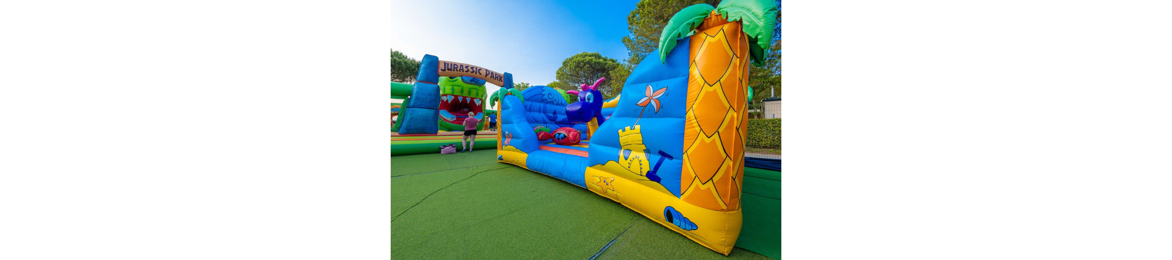 A bright inflatable play structure with cartoon graphics stands on a green outdoor surface. The design features a blue dragon, sandcastle, and palm tree illustrations in vivid colors. In the background, another inflatable attraction with a large dinosaur mouth and the text "JURASSIC PARK" is visible. A person walks nearby, suggesting a family-friendly amusement or festival setting.