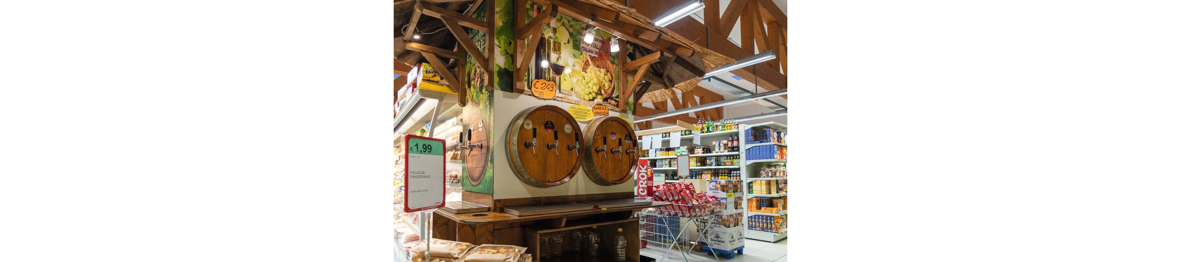 A supermarket interior features large wooden barrels with taps for dispensing beverages. Above the barrels, colorful signage shows grapes and a price tag reading €2,39. Nearby shelves are stocked with various packaged foods and drinks, and a shopping cart is visible in the aisle. A green and white sign on the left displays a price of 1,99 for a meat product.