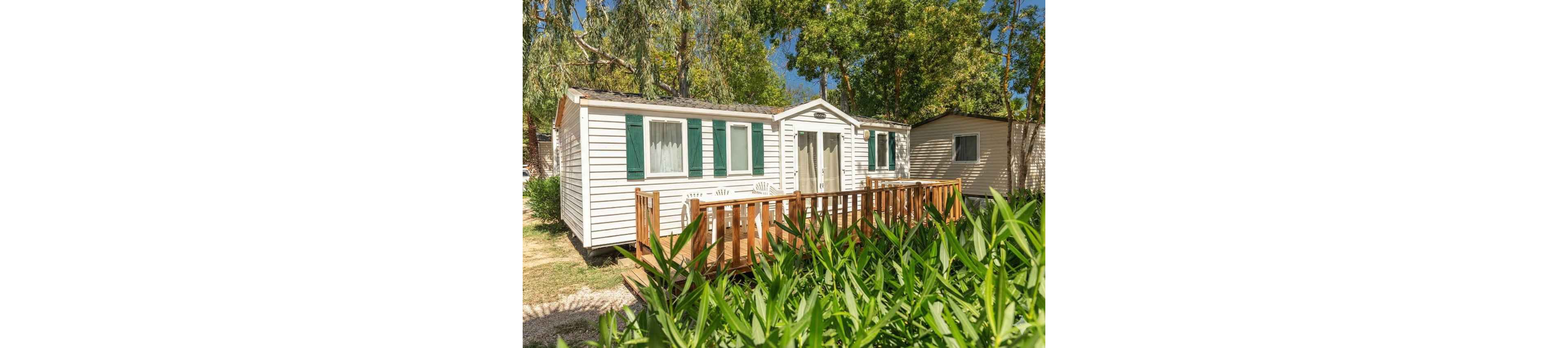 A white mobile home with green shutters stands in a sunny outdoor setting. The structure features a small wooden deck with railings at the front entrance. Lush green plants and trees surround the home, creating a natural, secluded atmosphere. Bright daylight and clear skies suggest a warm, pleasant day in a holiday park or campground.