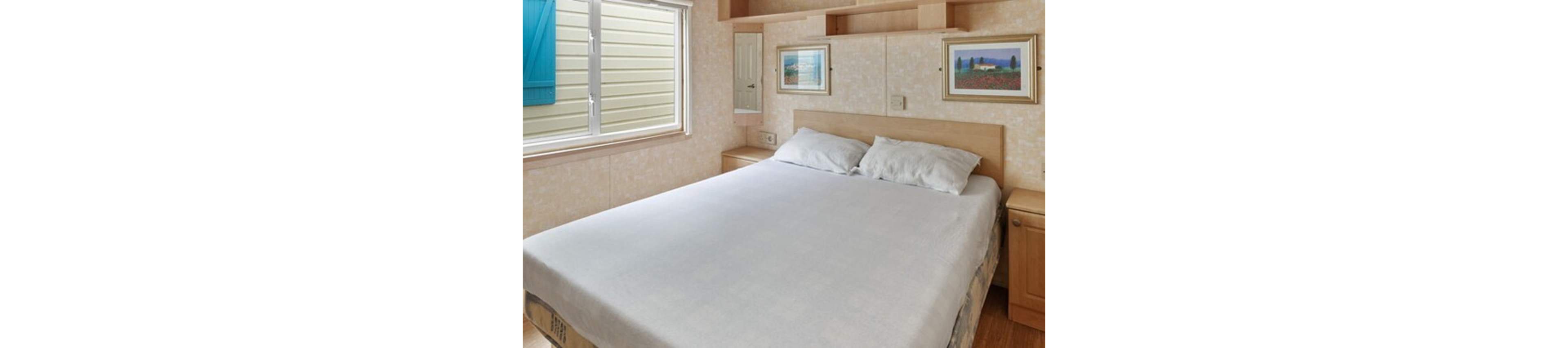A bright bedroom features a large neatly made bed with white bedding and two pillows. Light wood furniture surrounds the bed, including built-in shelves and cabinets. A window lets in natural light and shows the exterior siding of a neighboring structure. Two framed landscape pictures hang above the headboard, adding subtle color to the neutral room.