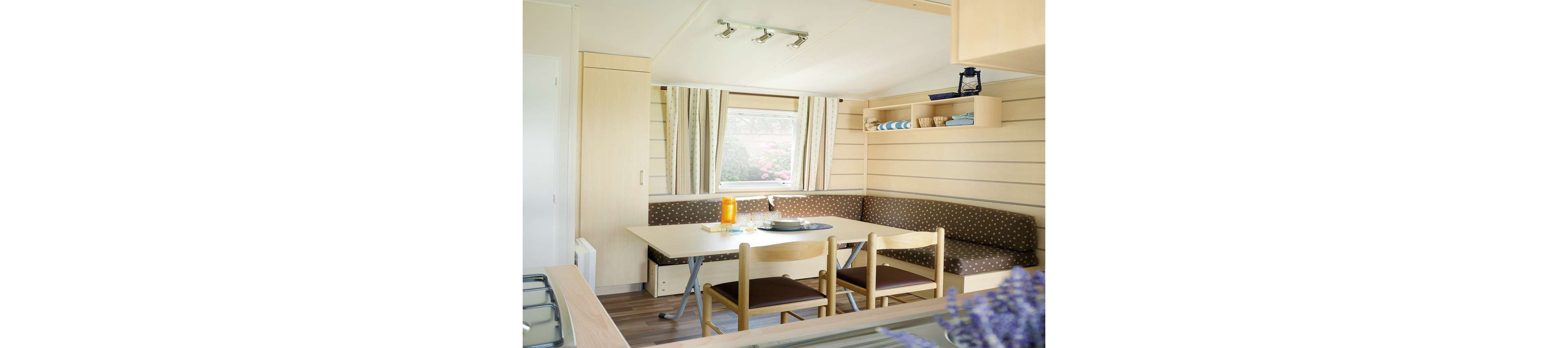 Cozy dining nook with a corner bench and wooden chairs is set inside a compact mobile home or cabin. A rectangular table holds a carafe of orange drink and glasses, positioned near a window with striped curtains. Light wood paneling, built-in shelving with folded towels, and spotlights on the ceiling create a clean, modern look. The foreground shows part of a kitchen counter and stove, emphasizing the small open-plan living space.