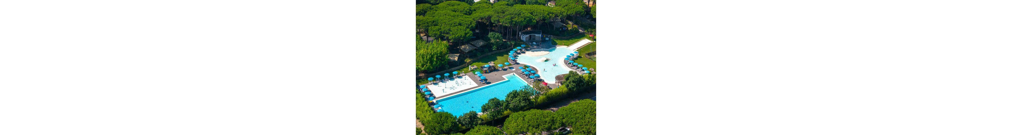 An aerial view shows a large resort-style swimming pool complex surrounded by lush green trees. The scene includes a rectangular lap pool and an irregularly shaped leisure pool with shallow areas and water features. Numerous blue umbrellas and sun loungers line the pool decks, suggesting a relaxing holiday atmosphere. Buildings and pathways are visible around the pools, indicating a well-developed recreational facility.
