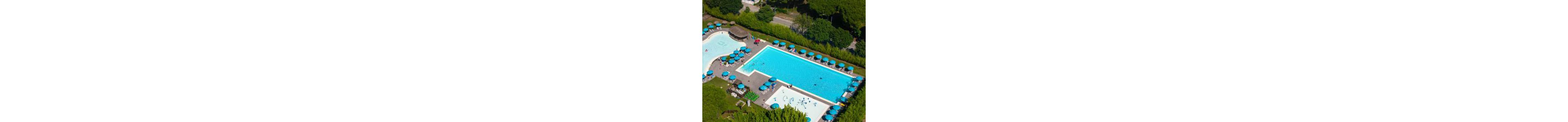 A large rectangular swimming pool complex is seen from above in a lush green resort setting. Multiple pools of different shapes are surrounded by rows of blue sun loungers and umbrellas. A few people relax in and around the water, creating a leisurely vacation atmosphere. Trees, hedges, and a nearby road frame the pool area, emphasizing its organized layout and outdoor environment.