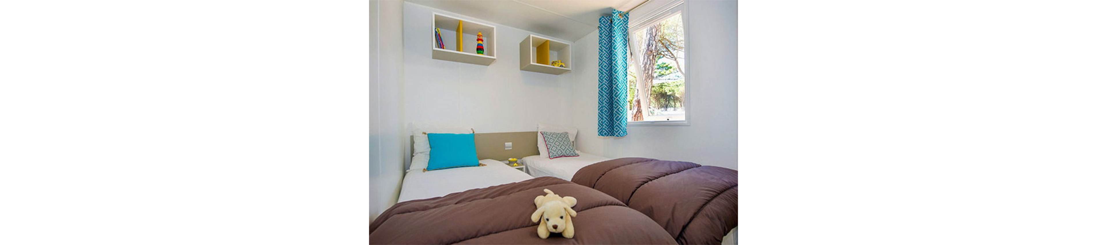 A cozy bedroom features two single beds with brown duvets and colorful accent pillows. A small plush dog toy rests on the nearest bed, adding a playful touch. Built-in wall shelves hold books and decorative items above the headboards. Natural light enters through a window with blue patterned curtains, revealing trees outside and creating a bright, inviting atmosphere.