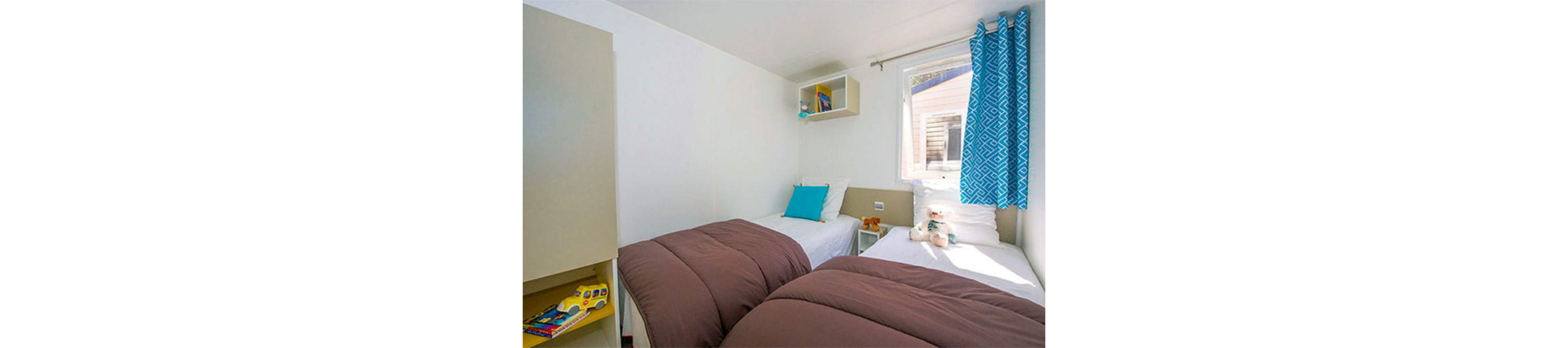 A compact bedroom features two single beds with brown duvets and white sheets. The room is bright, with natural light coming through a window covered by blue patterned curtains. A small nightstand between the beds holds a teddy bear and other toys, while shelves display books and a toy vehicle. The clean white walls and minimal decor create a simple, cozy atmosphere suitable for children or guests.