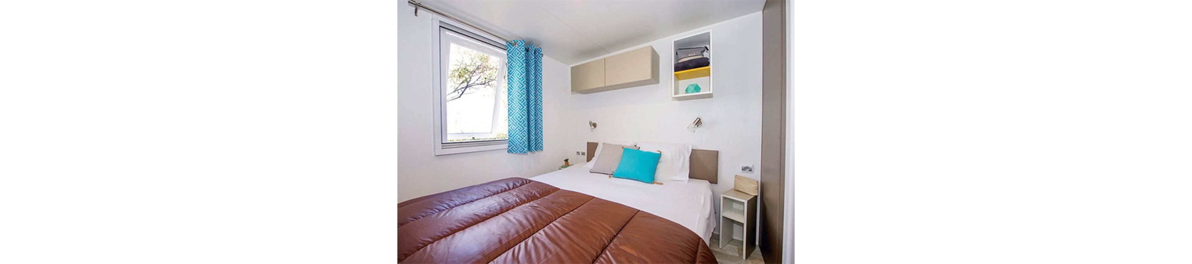 Compact modern bedroom featuring a neatly made double bed with a brown duvet and white sheets. Turquoise and beige throw pillows add color against a simple white wall. A window with patterned turquoise curtains lets in natural light and a glimpse of trees outside. Minimalist wall cabinets and open shelving provide storage, creating a clean and organized look.
