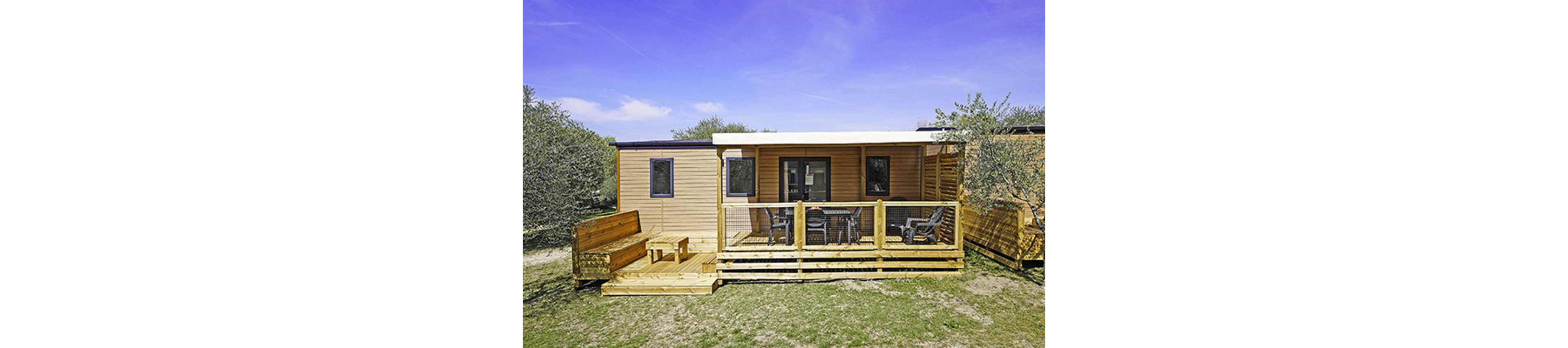 A contemporary wooden mobile home stands in an open outdoor setting under a bright blue sky. The structure features a covered front terrace with several chairs and a table, creating a comfortable seating area. A built-in wooden bench and table sit on an adjacent deck extension, enhancing the outdoor living space. Sparse grass and surrounding shrubs suggest a natural, campsite-like environment ideal for relaxation.