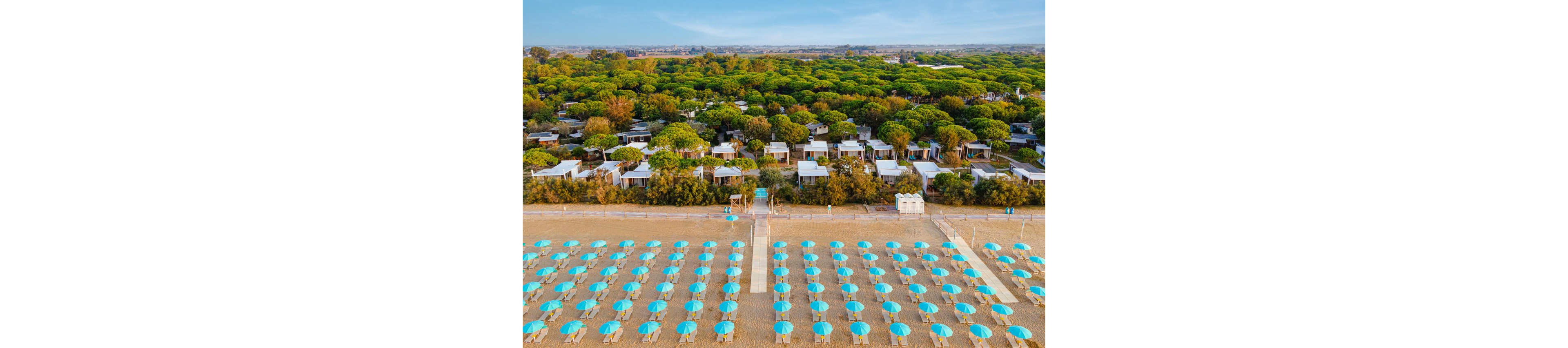 Aerial view of a sandy beach lined with rows of turquoise umbrellas and sun loungers. A central wooden walkway leads from the shoreline toward a cluster of small white resort cabins. Behind the cabins, dense green trees form a lush forested backdrop under a clear blue sky. The scene suggests a calm, well-maintained coastal holiday destination with strong geometric patterns.