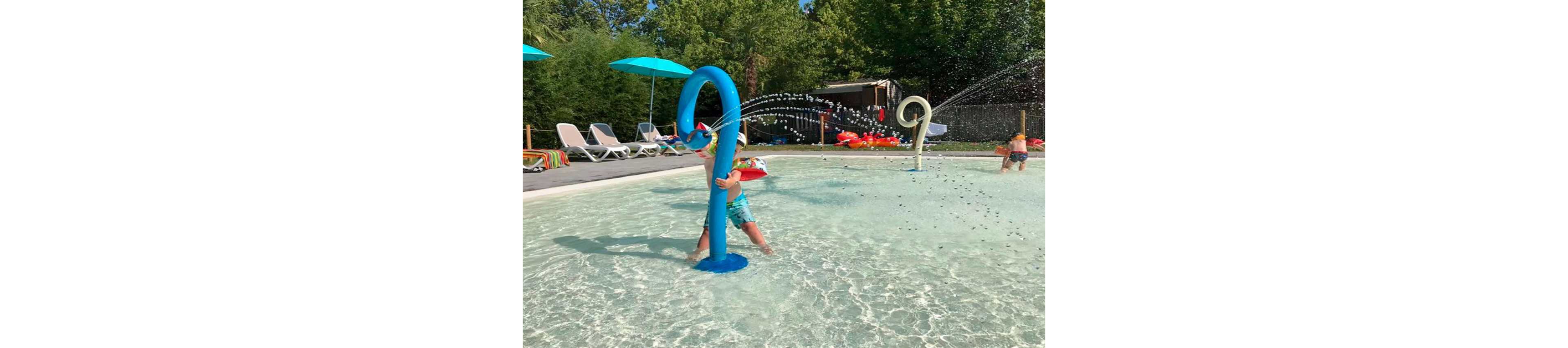 A young child plays with a curved blue water feature in a shallow splash pool. Water sprays in arcs from the structure, creating a playful fountain effect. Another child runs through the water in the background, while lounge chairs and umbrellas line the poolside. Trees and greenery surround the area, giving the scene a relaxed outdoor atmosphere.