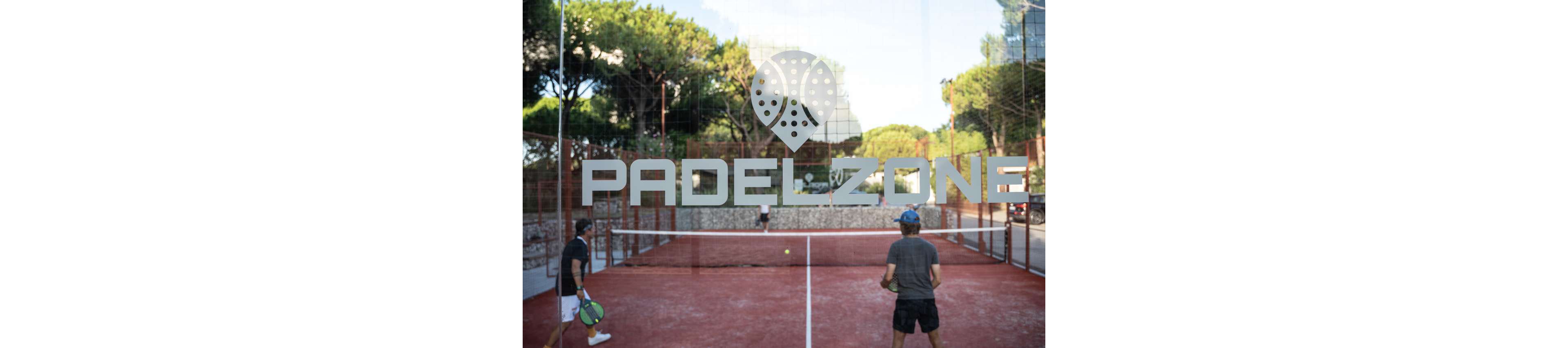 Two padel players stand on an outdoor red court, viewed through a glass wall. The word PADELZONE is prominently displayed on the glass above a stylized padel racket icon. Tall green trees and a stone wall frame the background, suggesting a club or sports facility setting. The scene captures a casual game in progress under natural daylight.