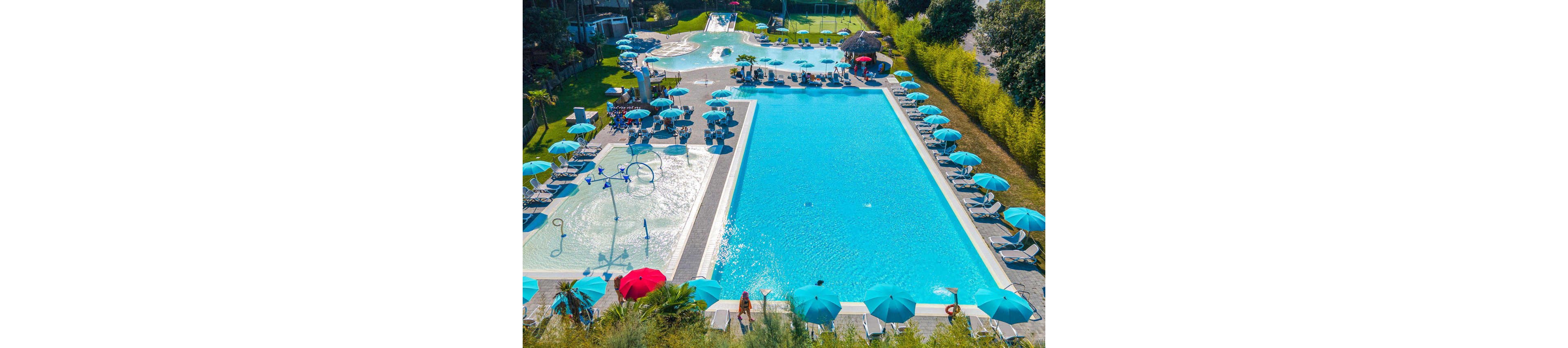 An aerial view shows a large rectangular swimming pool beside a shallower leisure pool at a resort. Numerous turquoise umbrellas and sun loungers line the pool decks, with people relaxing and swimming. A children’s splash area with water features is visible near one end, and a bar or shaded hut area appears in the background. Lush greenery and trees surround the complex, creating a tropical vacation atmosphere.