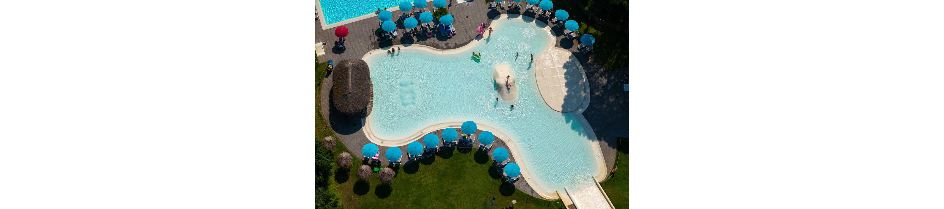 An aerial view shows a resort-style swimming pool with a unique organic shape. People relax on lounge chairs under bright blue umbrellas surrounding the pool. Children and adults enjoy the shallow water, central play area, and a wide slide leading into the pool. The scene is framed by green grass and landscaped trees, creating a vibrant vacation atmosphere.