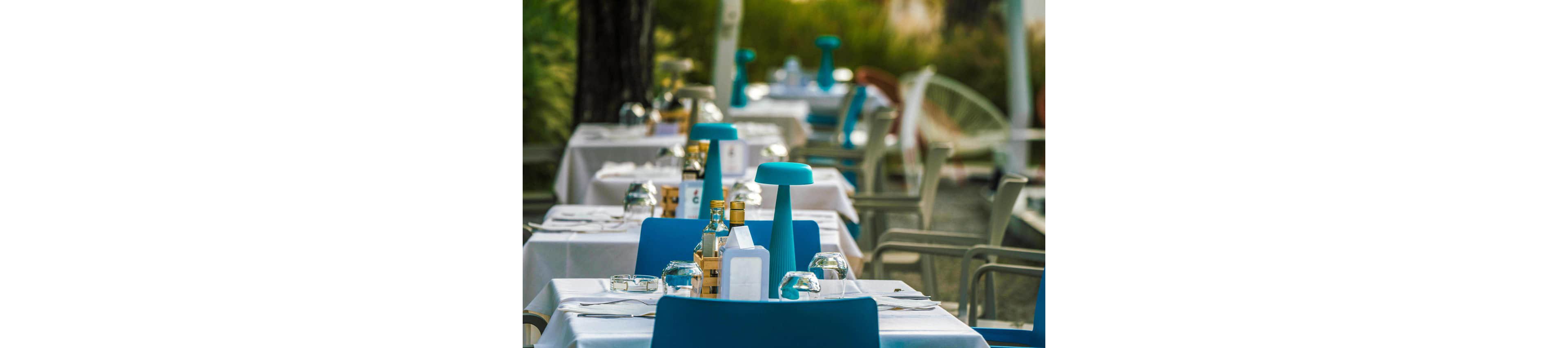 Neatly arranged outdoor restaurant tables are set with white tablecloths and bright blue chairs. Each table features a modern blue table lamp, glassware, and bottles of oil or condiments. The scene is empty of diners, suggesting preparation before service or a quiet moment between seatings. Soft greenery in the background adds a relaxed, summery atmosphere.