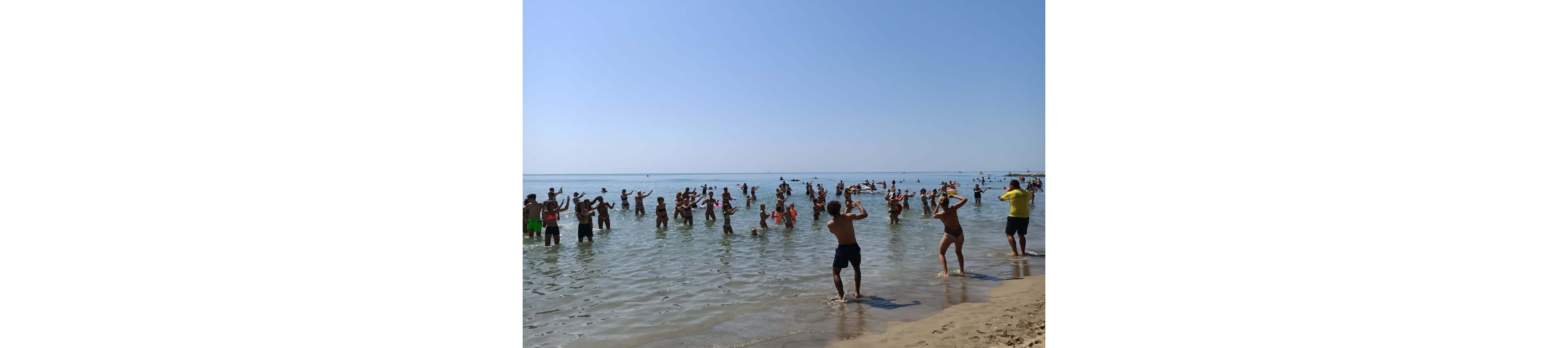 A large group of people stand in shallow seawater near a sandy beach, participating in a coordinated exercise or dance routine. The scene takes place on a clear, sunny day with calm water and a bright blue sky. Participants of various ages wear swimsuits and raise their arms, following the leader near the shore. The distant horizon and open sea emphasize a lively summer vacation atmosphere.