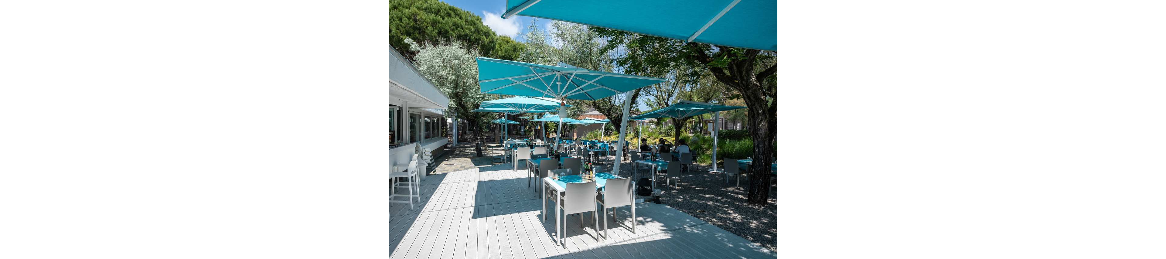 An open-air restaurant terrace features neatly arranged tables and chairs under large turquoise umbrellas. The setting is a modern dining area beside a building with large windows, surrounded by trees and greenery. Sunlight casts soft shadows across the light wooden deck, creating a relaxed and inviting atmosphere. A few people sit at tables in the background, suggesting a casual meal or coffee break in a peaceful outdoor environment.