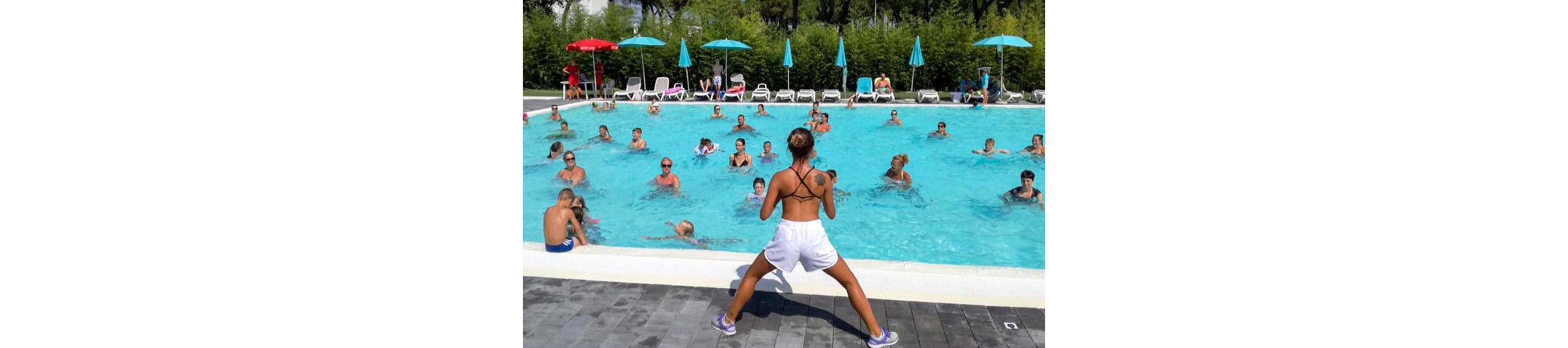 A fitness instructor stands at the edge of an outdoor swimming pool leading a large aqua aerobics class. Adults and children follow the exercises in the bright blue water under sunny weather. Colorful umbrellas, lounge chairs, and greenery surround the pool, creating a relaxed vacation atmosphere. The scene suggests a lively group workout at a resort or public pool.