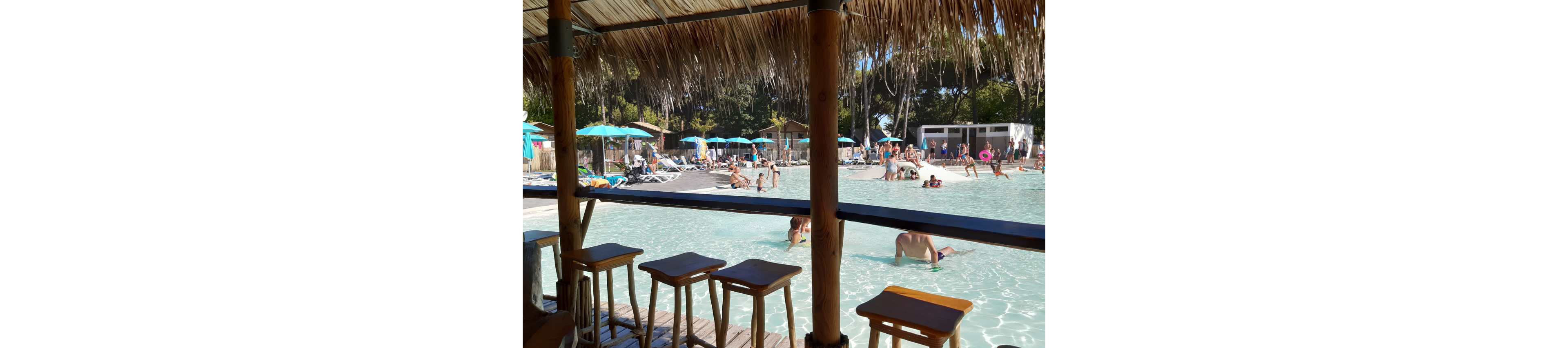 A shallow resort swimming pool is seen from under a thatched-roof bar area with wooden stools in the foreground. Families and children play in the clear water while others relax on lounge chairs around the pool. Colorful umbrellas provide shade along the poolside, and trees and buildings frame the background. The bright sunlight and turquoise water create a relaxed, vacation atmosphere.